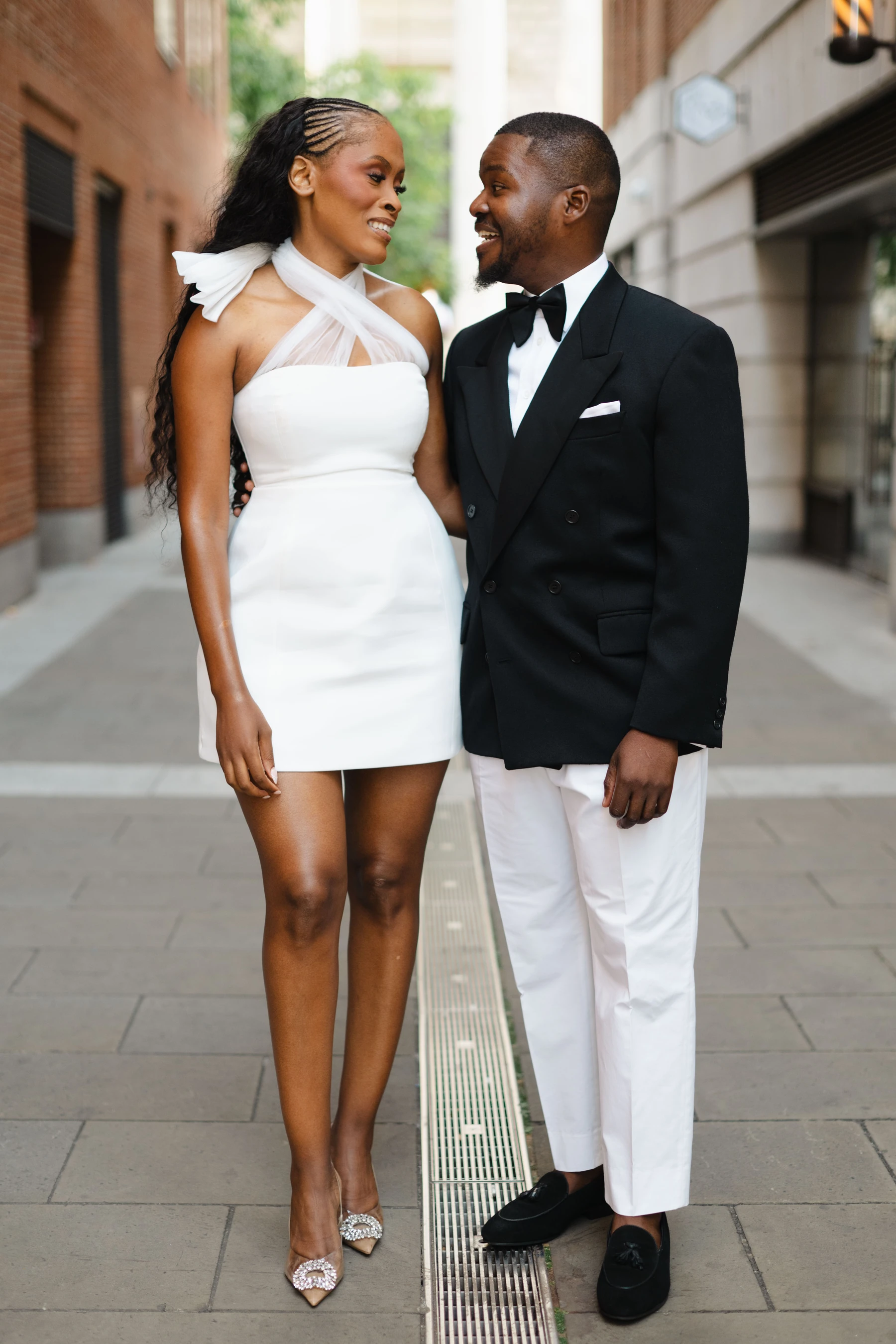 Kennedy and Didimalang smiling at each other on a city street, she in a white dress and he in a black tuxedo jacket